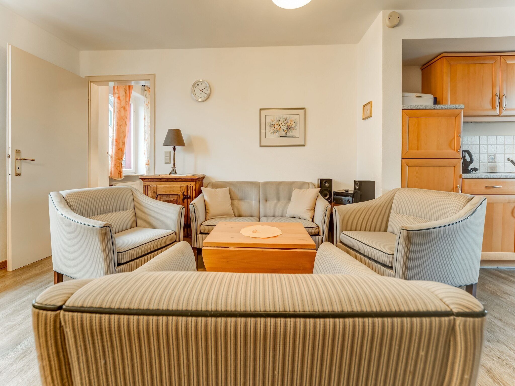 Ein Wohnzimmer mit gestreiften Sesseln, einem Sofa, einem Couchtisch, einer Uhr, einer Lampe und einer Küchenzeile mit Holzausbau.