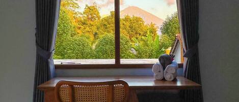 A room with a wooden desk and chair, a window with curtains, and a view of a mountain and greenery.