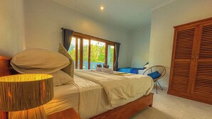 A bedroom with a bed, a bedside lamp, a chair, a wardrobe, and a window with curtains. - Rumah Diyah (Karangasem)