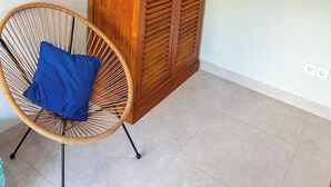 A tall wooden wardrobe with louvered doors in a room with a tiled floor and a wicker chair with a blue cushion. - Rumah Diyah (Karangasem)