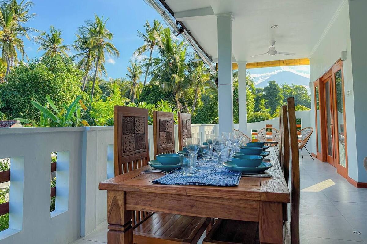 A dining area with a wooden table set for four, surrounded by chairs, with a view of palm trees and a mountain in the background.
