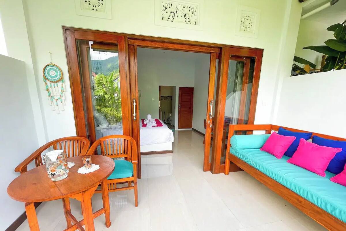 A room with a wooden table and chairs, a sofa with colorful cushions, and a sliding glass door leading to a balcony.