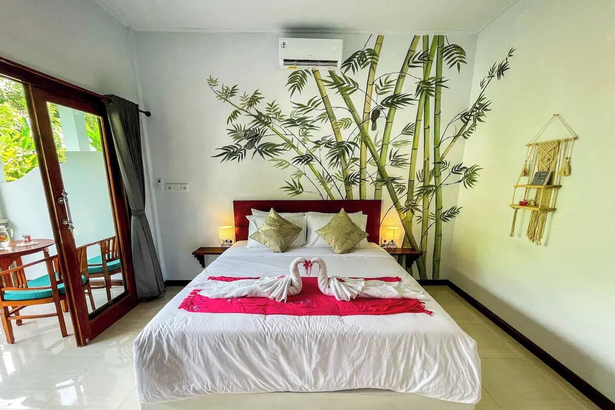 A hotel room with a bed, a large bamboo plant, a wall-mounted air conditioner, and a small table with a chair.