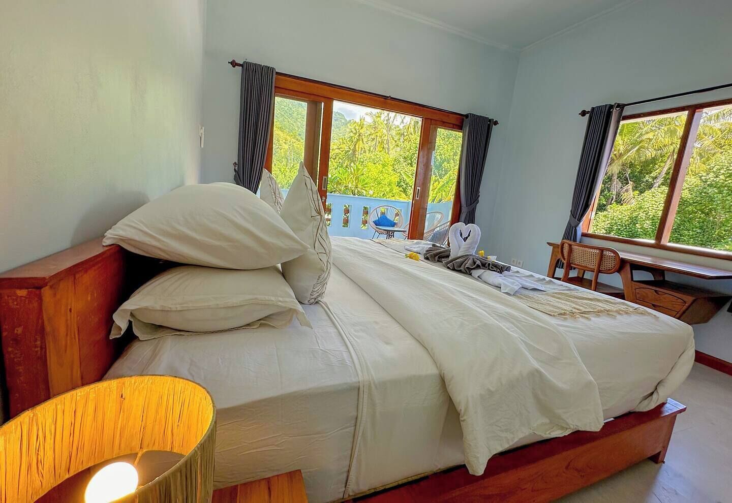 Un dormitorio con una cama grande, cabecera de madera y un escritorio con silla. Hay ventanas con cortinas y vistas a la vegetación exterior.