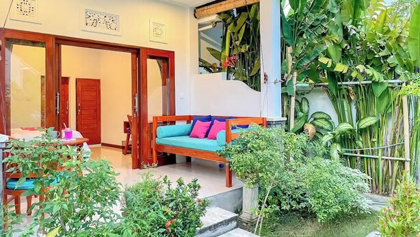 A cozy outdoor seating area with a wooden bench and cushions, surrounded by lush greenery and a staircase leading to an upper level. - Rumah Diyah (Karangasem)