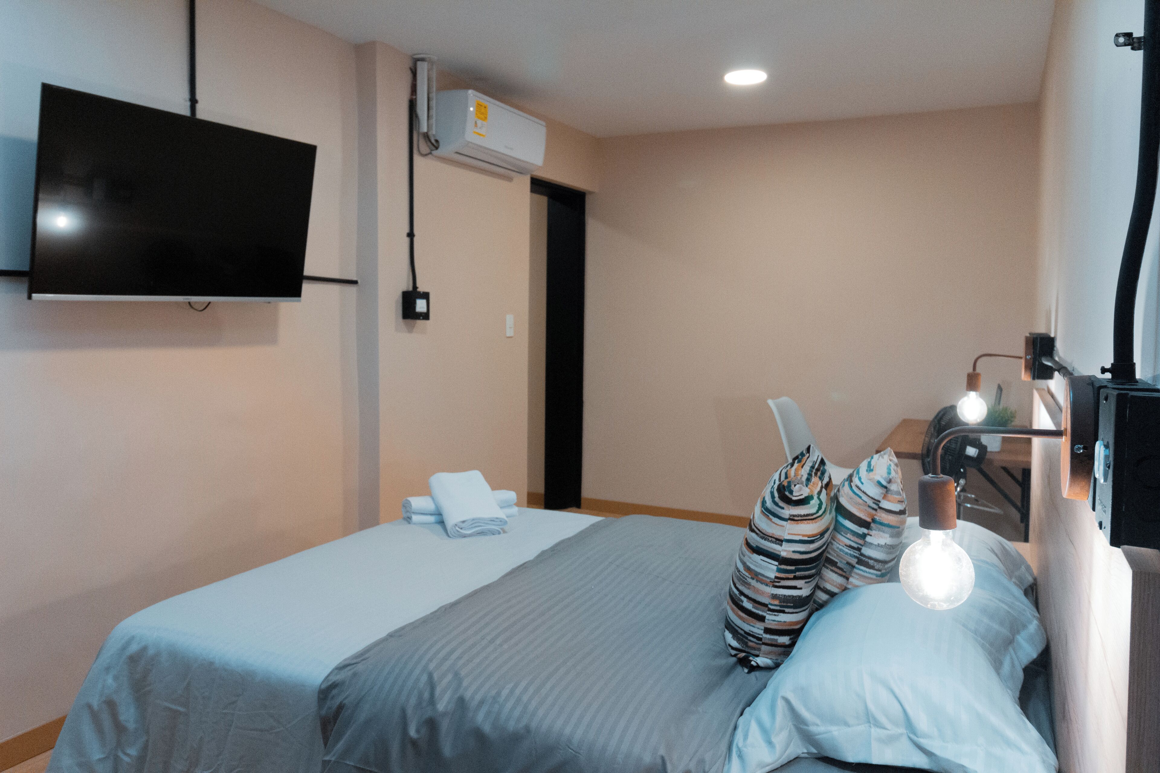 A hotel room with a bed, a TV mounted on the wall, and a bedside table with a lamp.