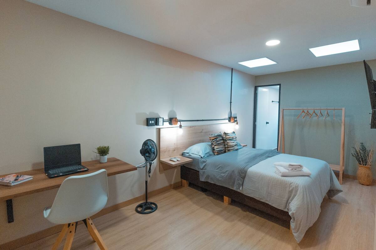 Une chambre à coucher avec un lit, un bureau, une chaise, un ordinateur portable, un ventilateur et une petite plante.
