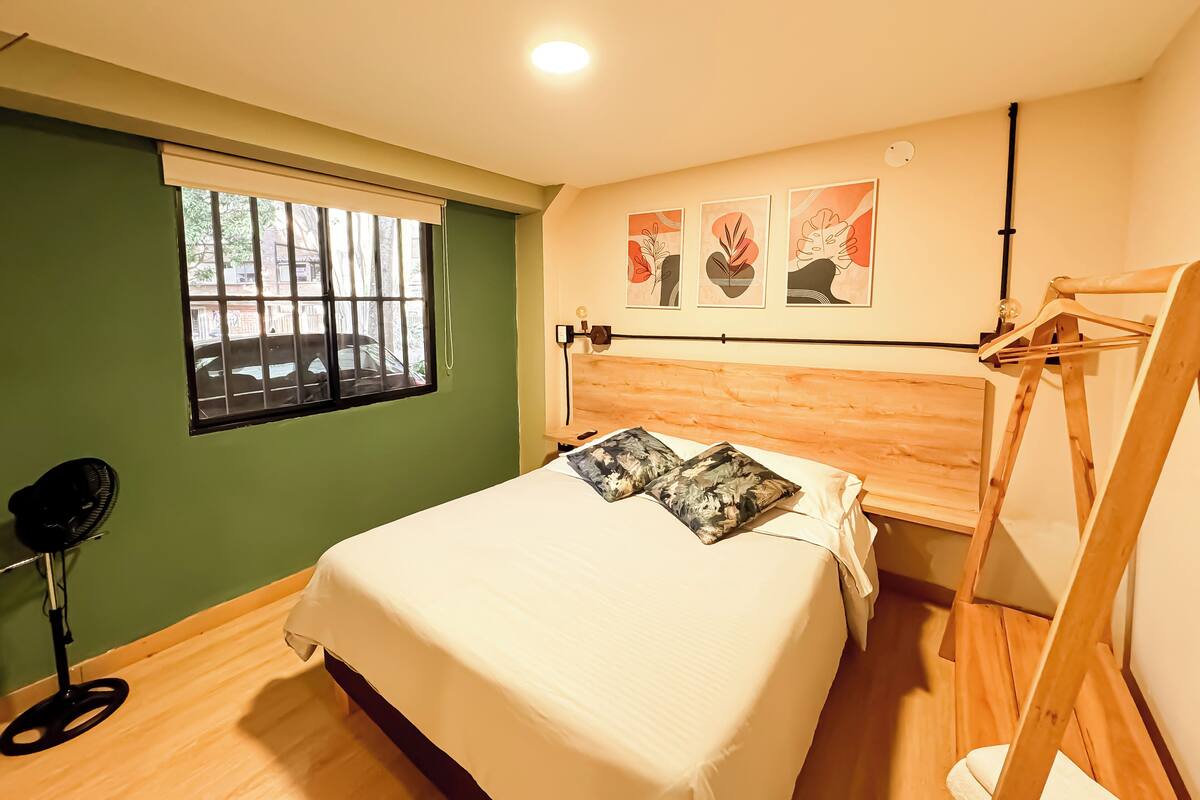 Une chambre à coucher avec un lit, un ventilateur sur pied, une fenêtre et des œuvres d’art accrochées au mur.