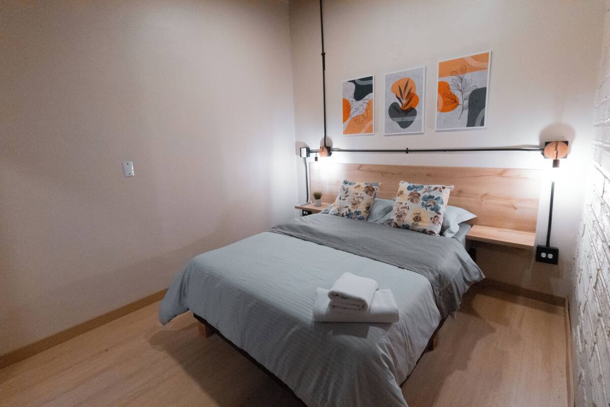 Un lit bien fait, avec une couvre-lit grise et des oreillers, une table de chevet avec une lampe, et trois œuvres d’art accrochées au mur.
