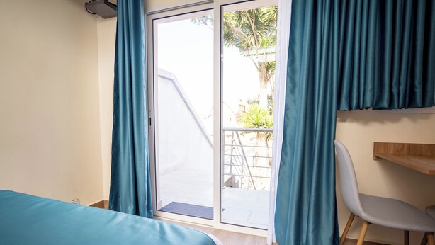 A room with a bed, a desk, and a chair. There is a sliding glass door leading to an outdoor area with a staircase and greenery. - Aparthotel Madeleine (Antananarivo)
