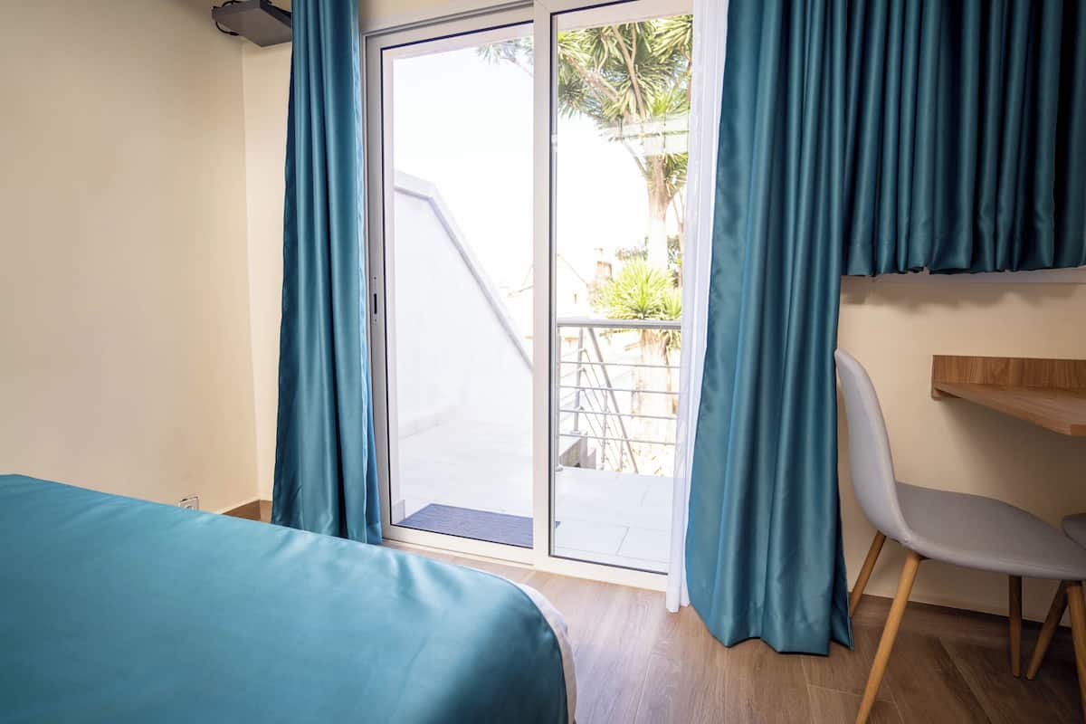 Una habitación con una cama, un escritorio y una silla. Hay una puerta corrediza de vidrio que da a un espacio exterior con una escalera y vegetación.