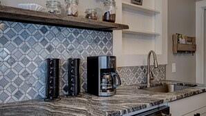 Une cuisine dotée d’un plan de travail en granit, d’une machine à café et d’un évier. Des étagères se trouvent au-dessus du plan de travail, où sont disposés des bocaux et un cadre photo.