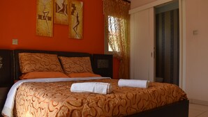 A bedroom with a bed, orange patterned bedspread, two pillows, a white towel, and artwork on the wall. - The Brilliant Villa Pernera (Pernera)
