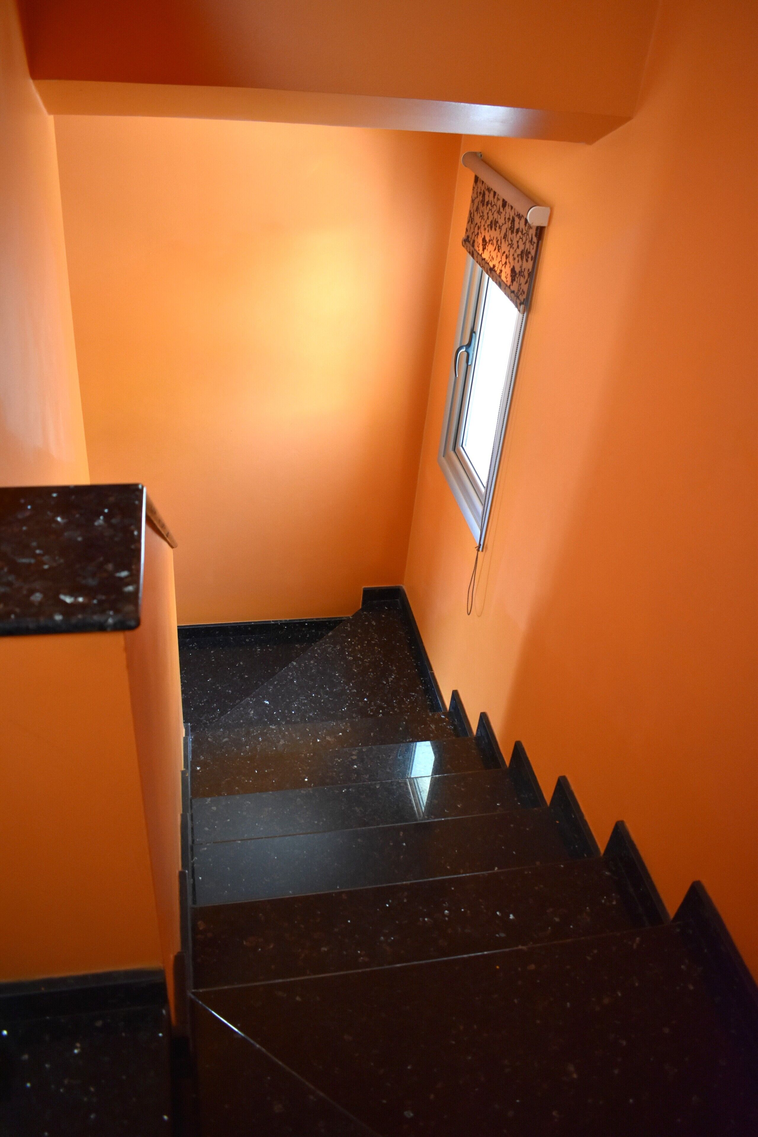 A staircase with black steps and orange walls, leading to a window with a patterned curtain.