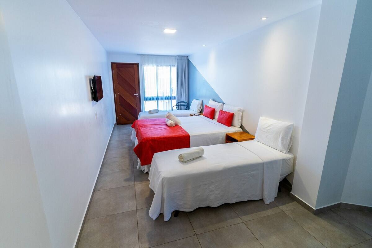 A hotel room with two beds, one with red and white bedding and the other with white bedding. There is a wooden door, a window with curtains, and a wall-mounted TV.
