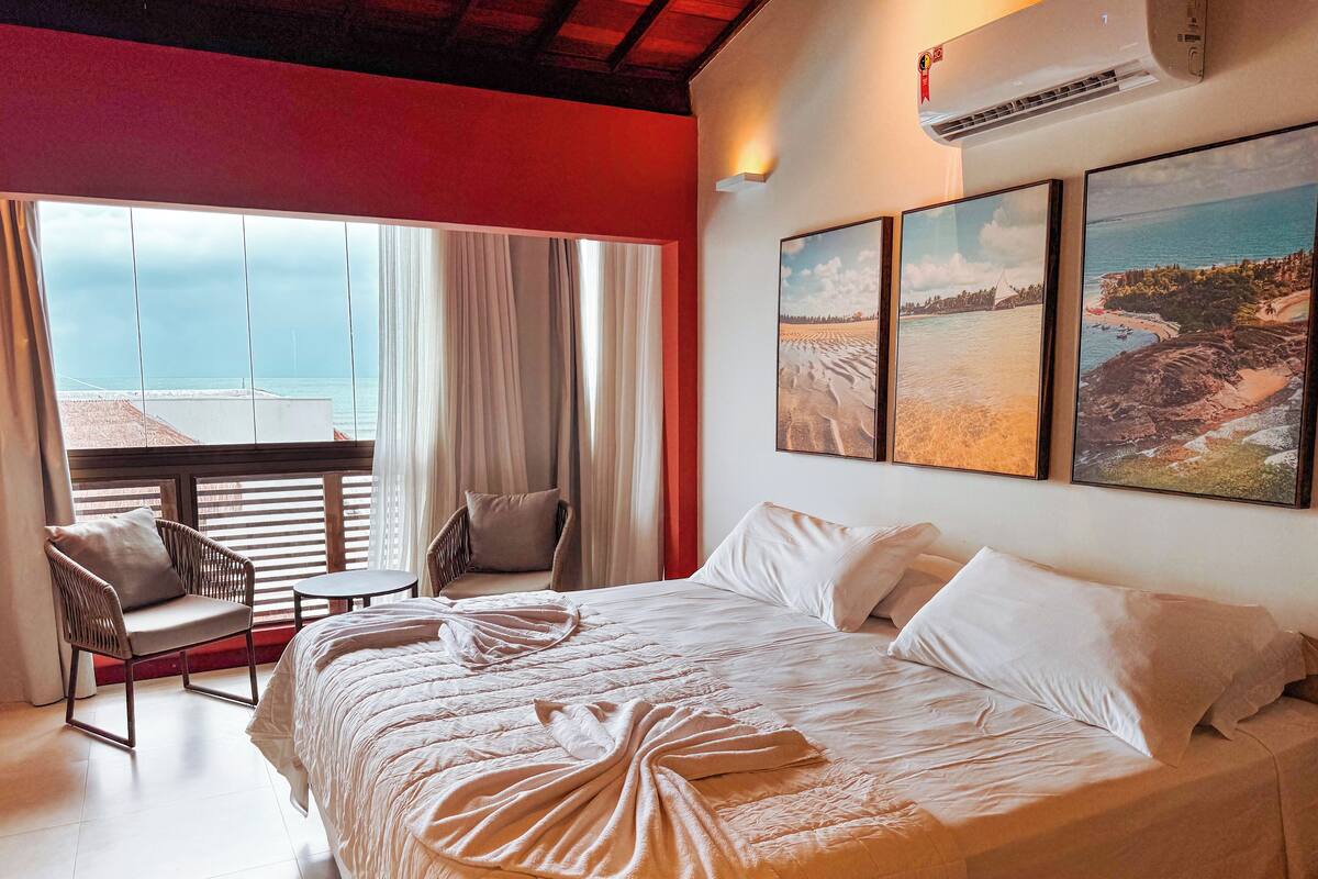 A hotel room with a large bed, a chair, a small table, and a view of the beach through the window.