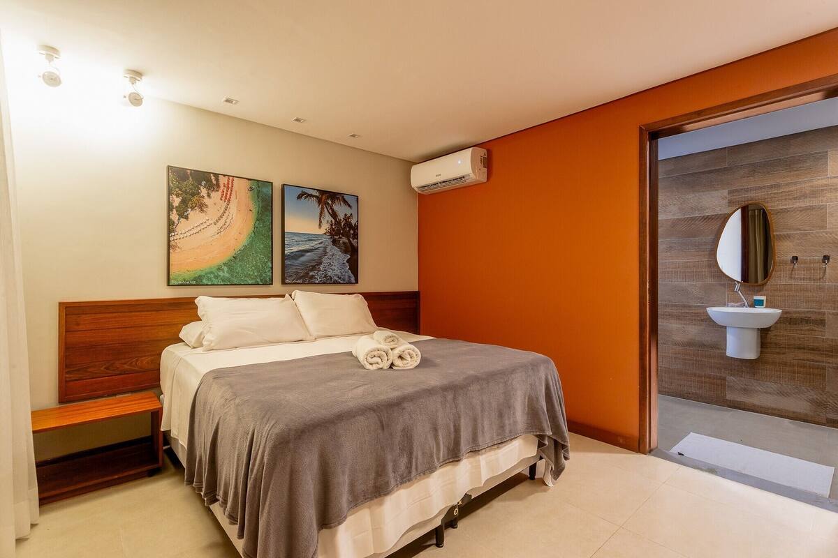 A bedroom with a bed, a bedside table, a wall-mounted air conditioner, and a bathroom with a sink and mirror.