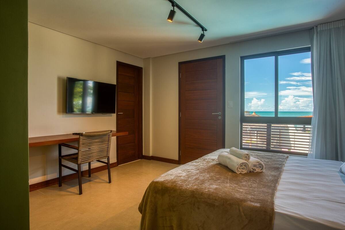 A hotel room with a bed, a desk, a chair, a TV, and a balcony with a view of the sea.
