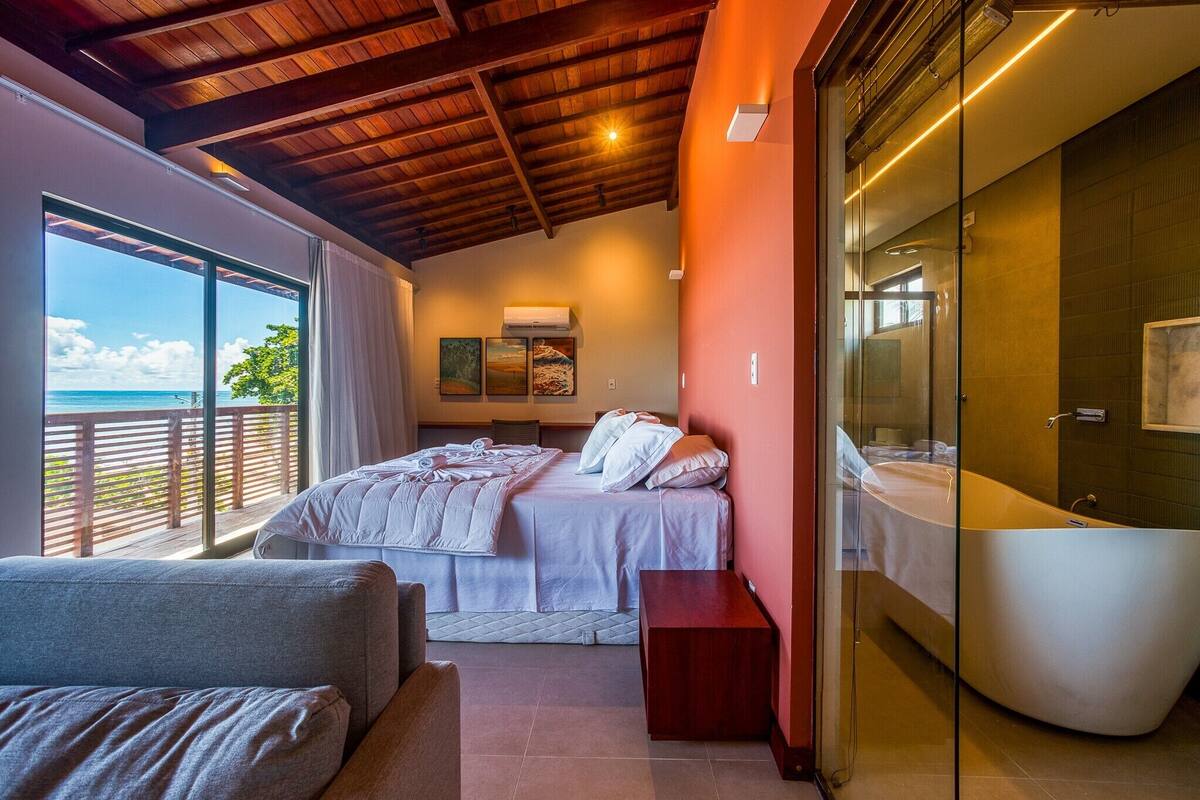 A bedroom with a large bed, a sitting area, a bathtub, and a balcony with a view of the ocean.