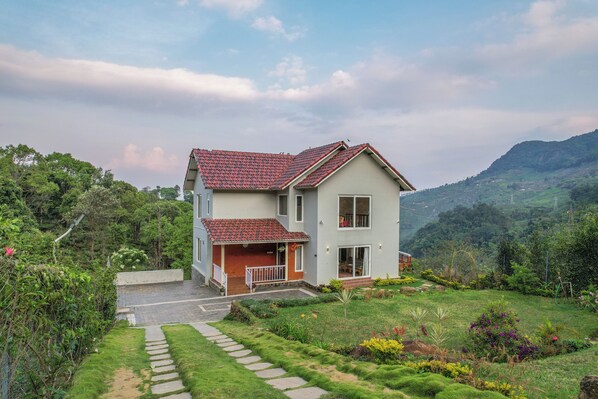 A two-story house with a red-tiled roof, surrounded by lush greenery and a mountainous backdrop. - Moving Mist Villa (Coonoor)