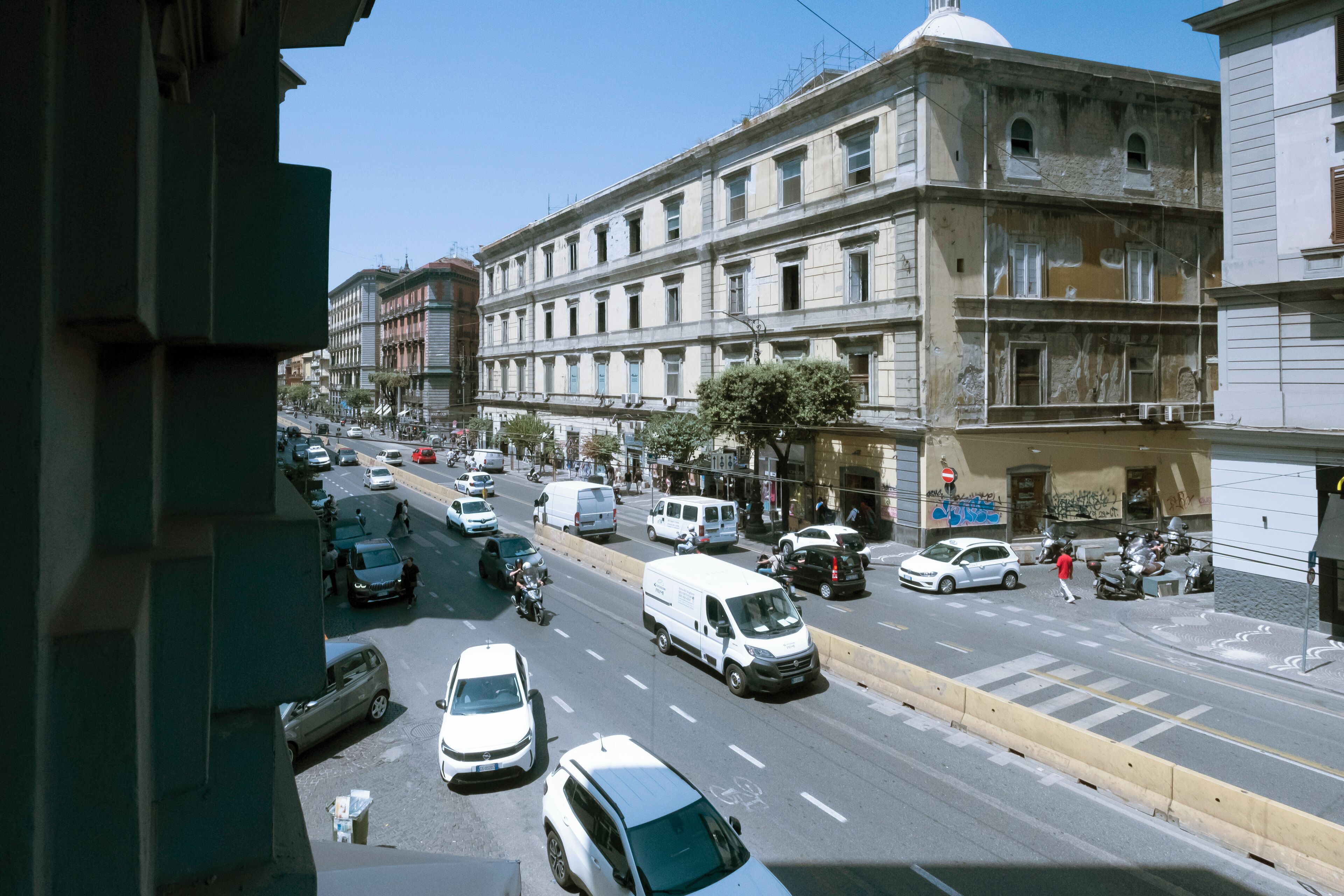 A busy urban street with multiple lanes of traffic, including cars, vans, and motorcycles. There are buildings with classical architecture, some with visible wear and graffiti.