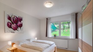 A bedroom with a wooden bed, a nightstand, a bedside lamp, a window with a view of a garden, and a painting on the wall. - Apartment with private terrace in Velmede (Bestwig-Velmede)
