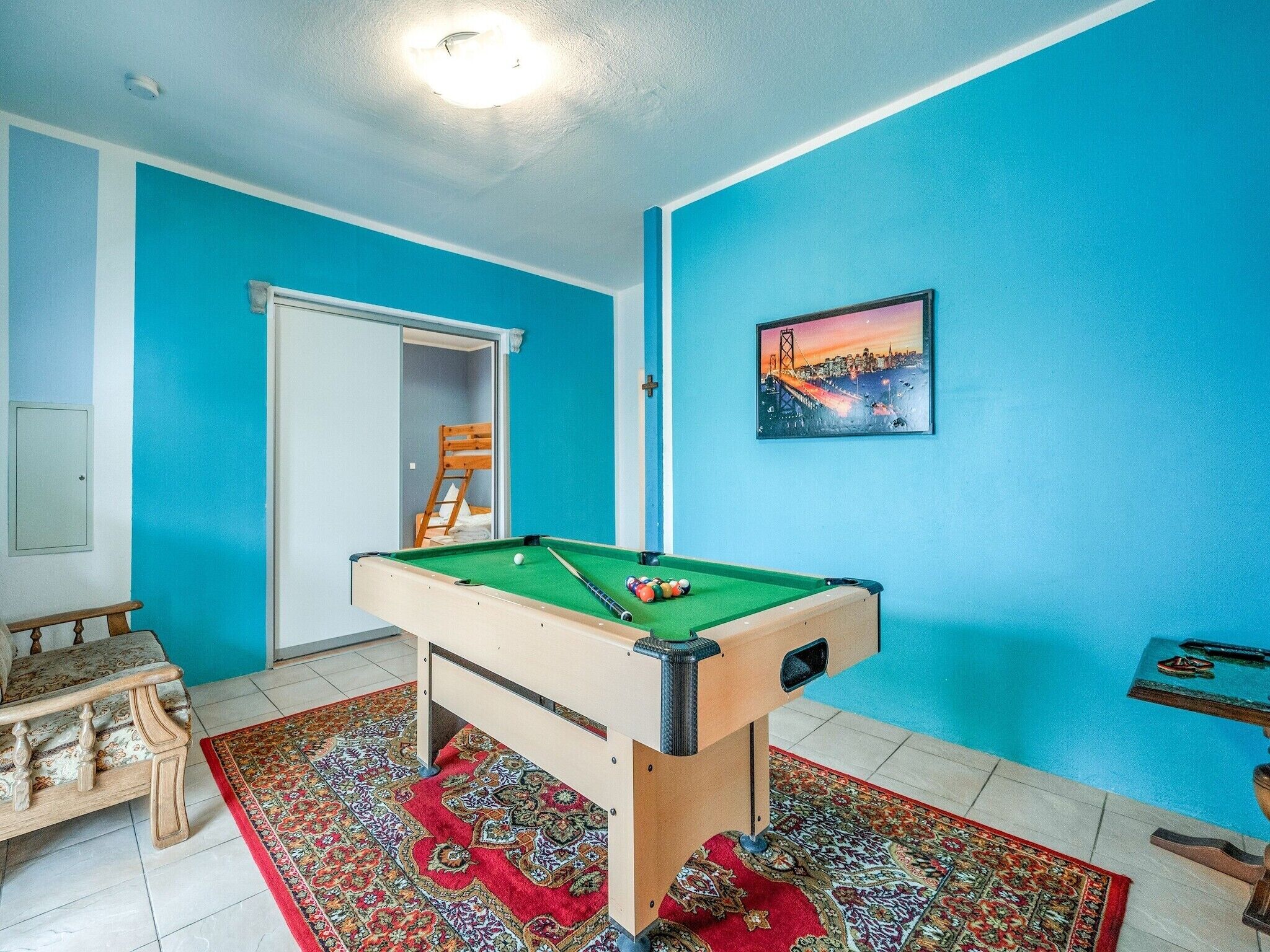 Une pièce comprenant une table de billard, un banc en bois, un tapis rouge à motifs et un tableau représentant un paysage urbain.