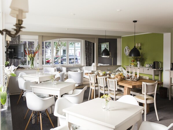 A modern dining area with white furniture, a wooden table, and a view of the street through large windows. - Winterberg Gruppenhaus mit Sauna – ideal für Familien & Freunde (Winterberg)