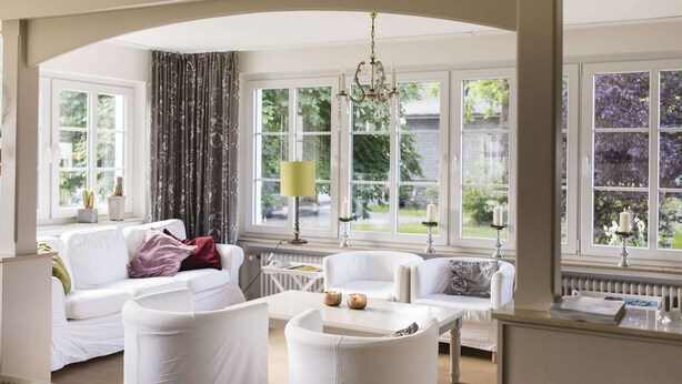 A spacious living room with white furniture, large windows, and a chandelier.