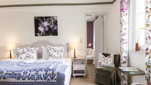 A bedroom with a bed, bedside tables, a nightstand, a chair, a small table, a lamp, a picture on the wall, and a window with curtains.