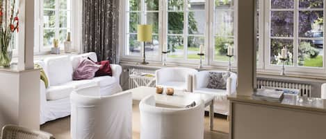 A spacious living room with a white sofa, armchairs, and a coffee table. Large windows provide a view of the garden.