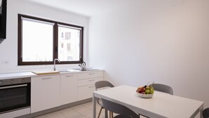 Une cuisine moderne avec des armoires blanches, un four encastré, un évier et une fenêtre au-dessus du plan de travail. On y trouve une table à manger sur laquelle est posé un panier de fruits.