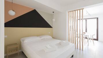 Habitació d'hotel moderna amb un llit gran, una taula de nit de fusta i una zona de menjador amb caires blancs.