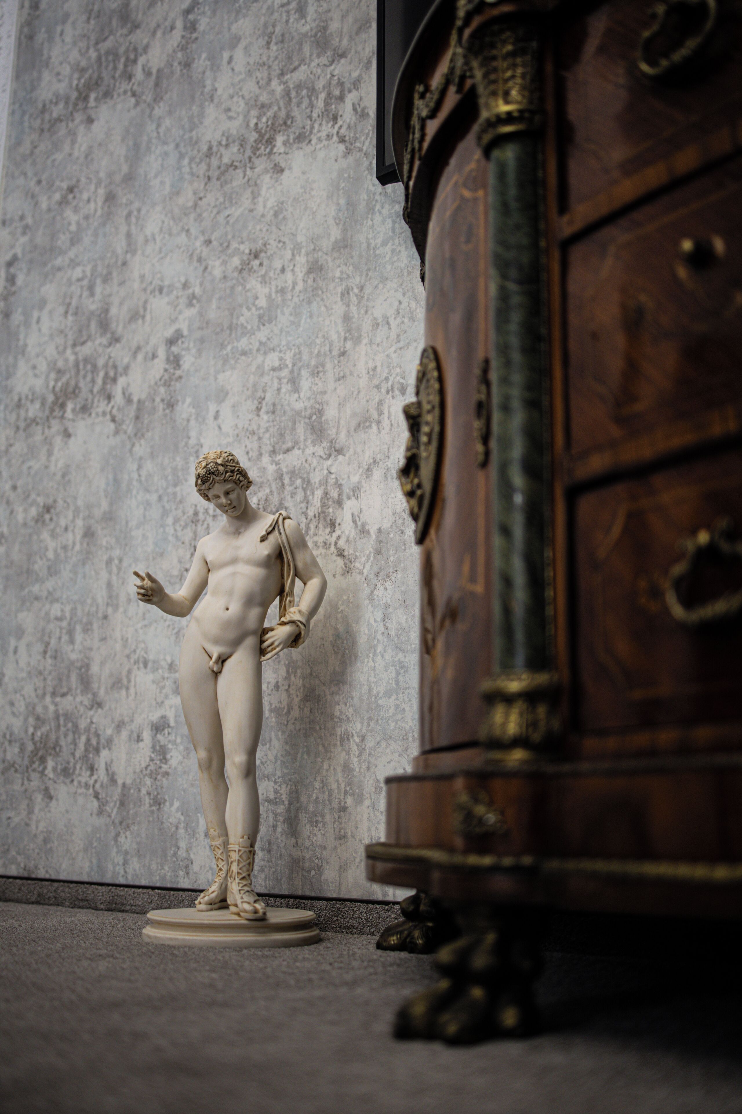Una estatua de mármol de una figura clásica sobre un pedestal, ubicada frente a una pared con textura, y un mueble de madera a la derecha.