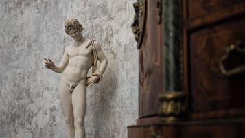 Una estatua de mármol de una figura clásica sobre un pedestal, ubicada frente a una pared con textura, y un mueble de madera a la derecha.