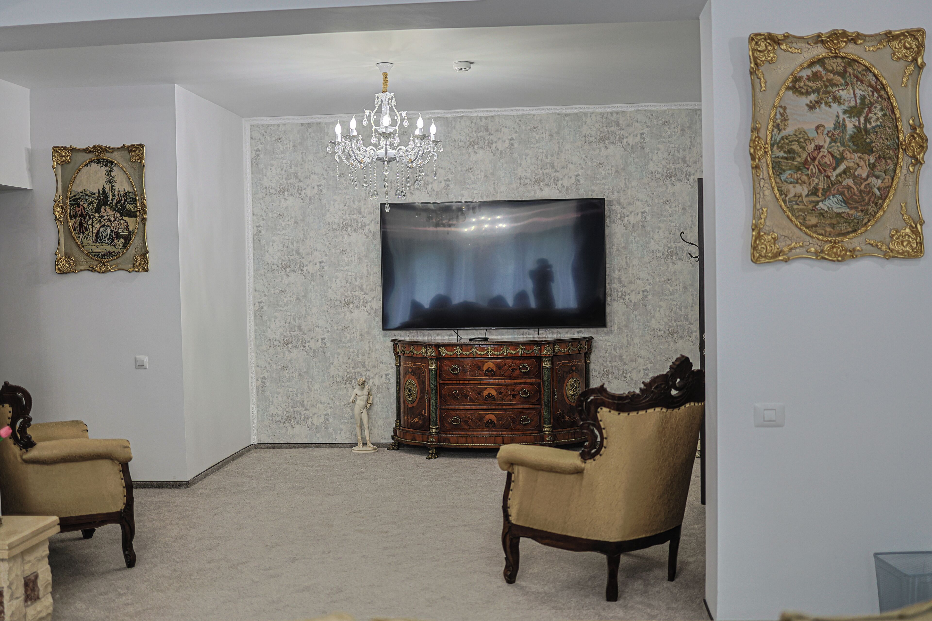 A living room with a large flat-screen TV, a chandelier, two armchairs, a wooden cabinet, and two framed artworks on the walls.