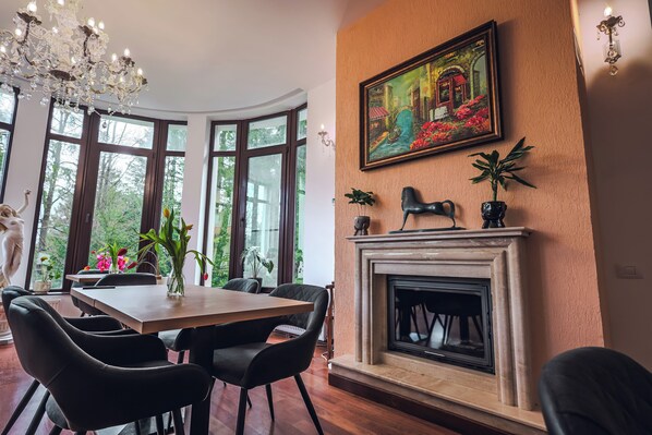 Un coin repas avec une table en bois et des chaises noires, une cheminée et une grande fenêtre donnant sur un paysage verdoyant.