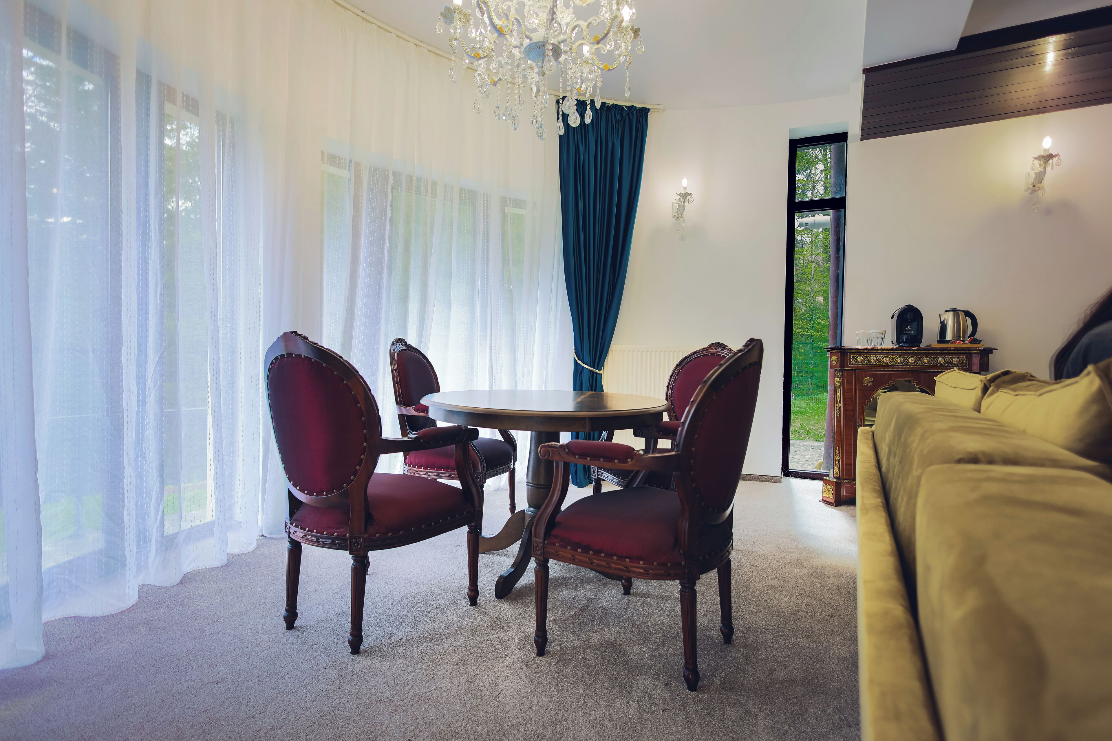 Ein Zimmer mit rundem Tisch, vier Stühlen, einer Chandelierleuchtre, einem Sofa und Blick auf Grün.