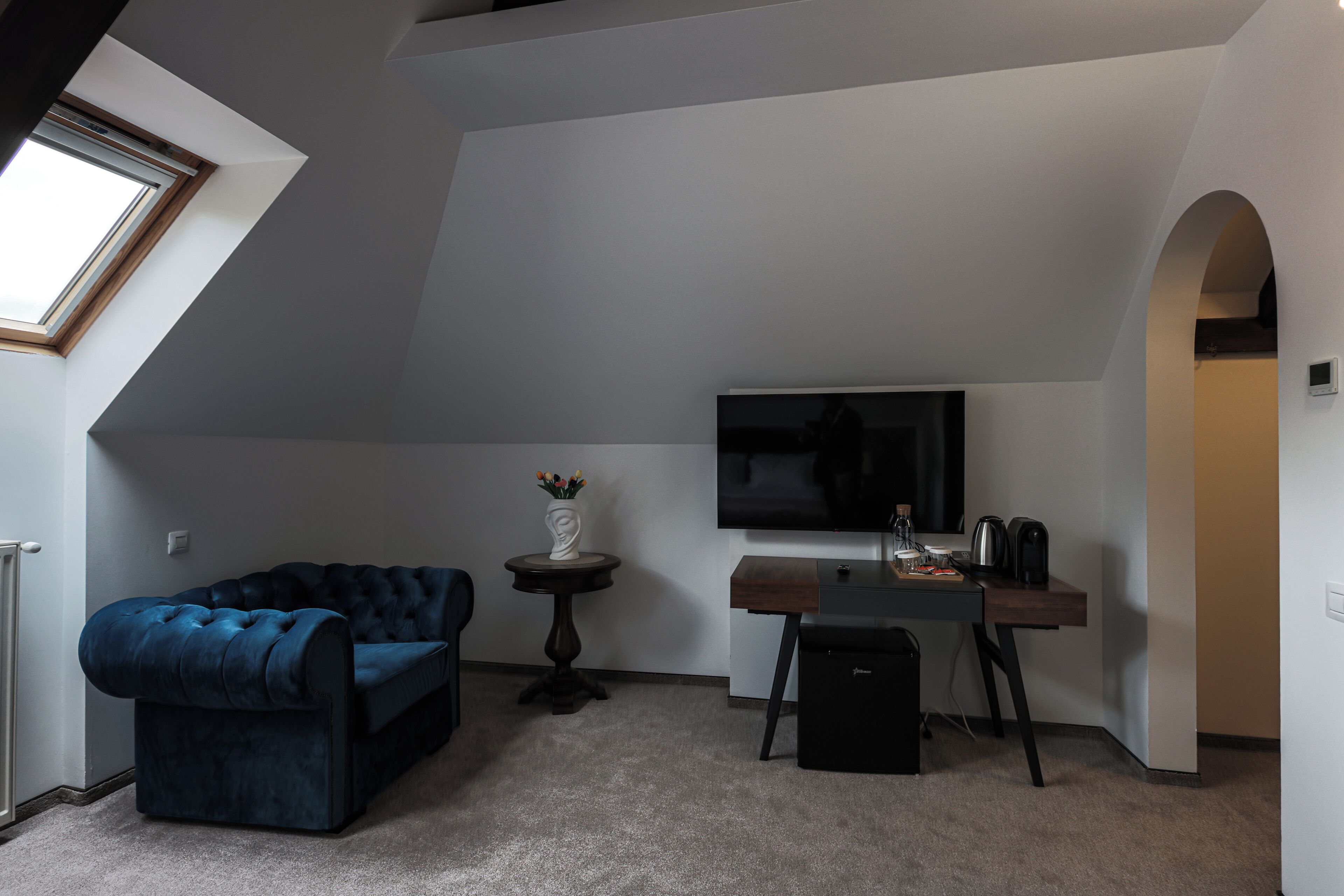A compact living space with a dark blue sofa, a small round table with a vase, a flat-screen TV, a coffee maker, and a desk with a computer.
