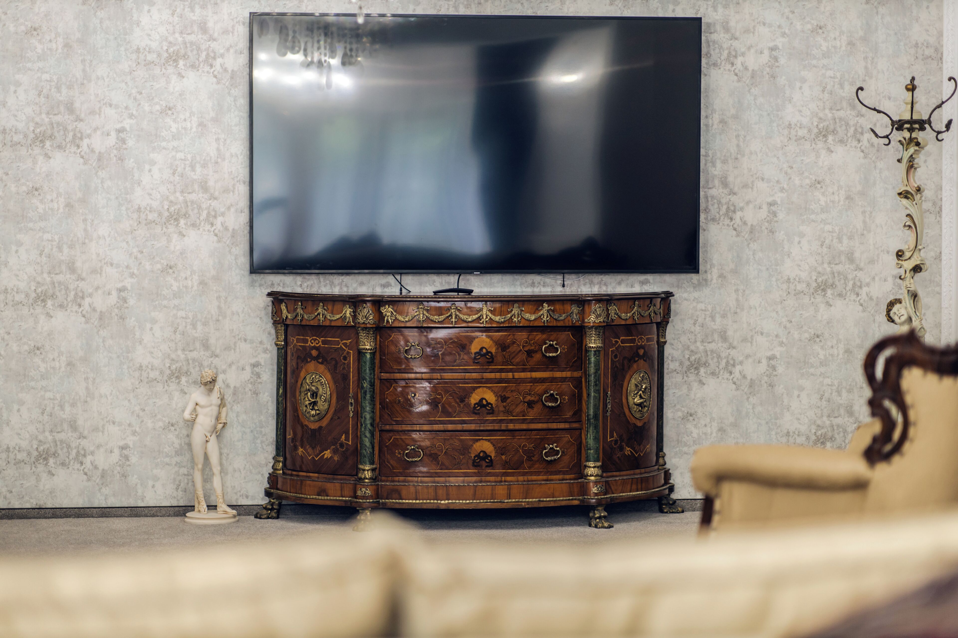 A room with a large flat-screen TV mounted on the wall, a wooden cabinet with drawers, a statue, and a chair.