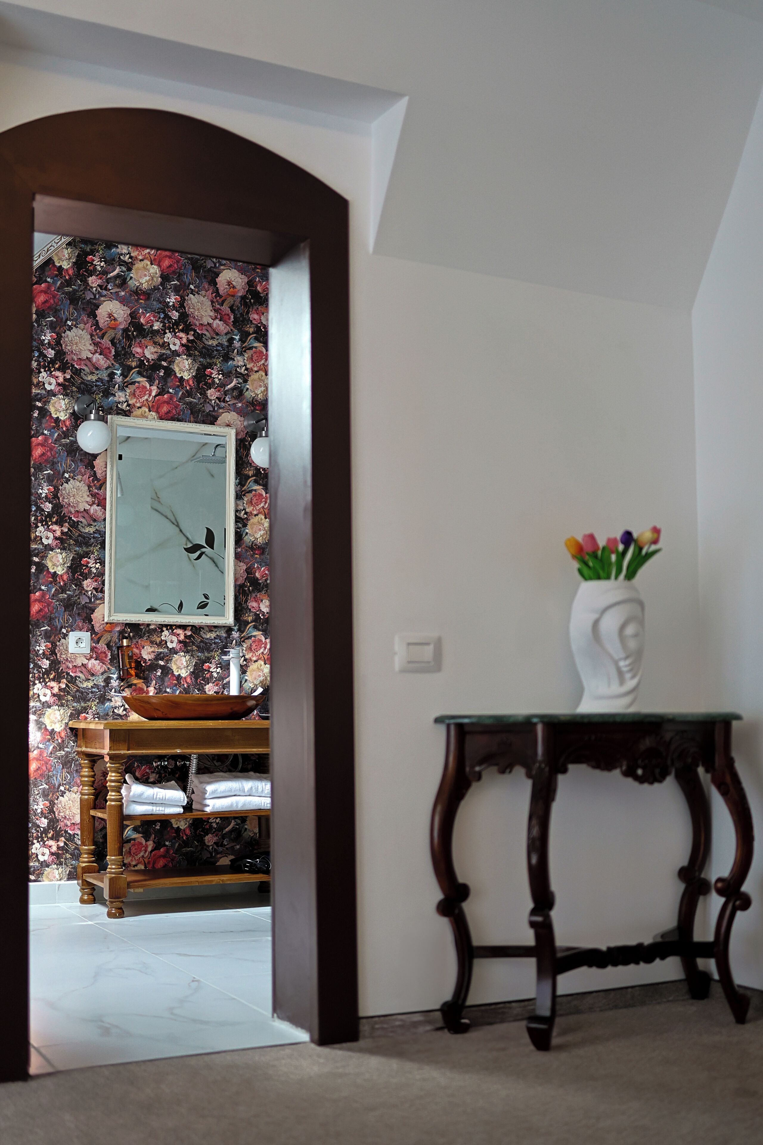 Ein Flur mit floralem Wandpapier, einem gerahmten Bild an der Wand, einer Holzbank, einer weißen Vase mit Blumen auf einem Tisch und einem Holzkasten.