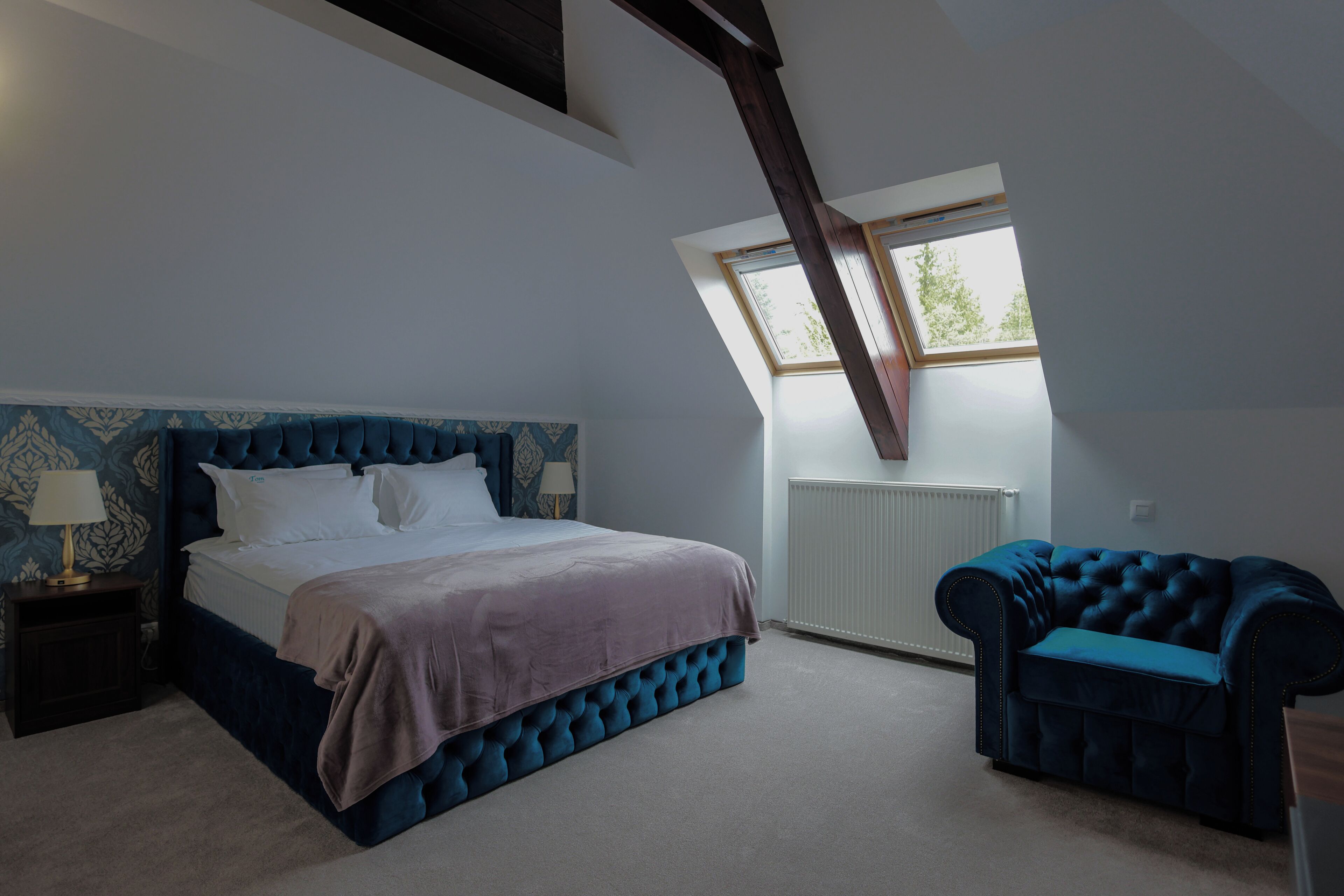 A bedroom with a large bed, a blue upholstered chair, a nightstand, and a window with a view of trees.