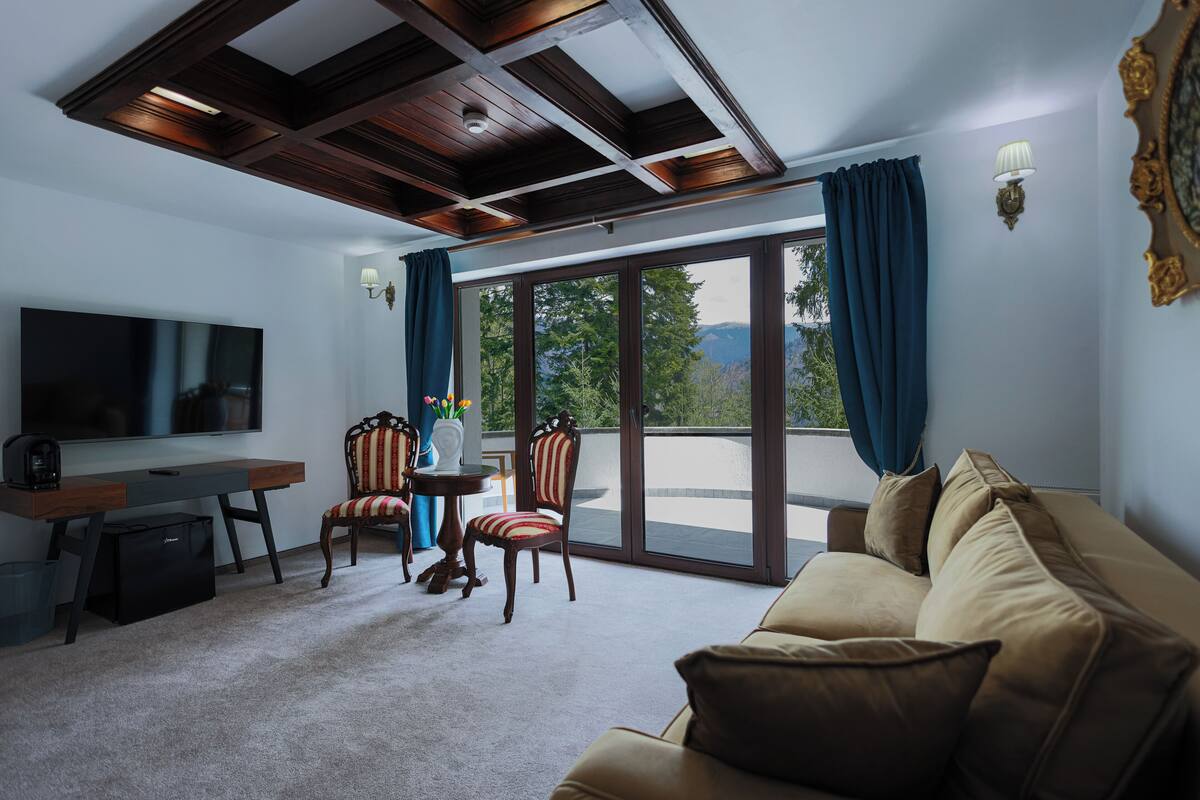 A living room with a sofa, a TV, a desk, and a chair. There is a balcony with a view of trees and mountains.