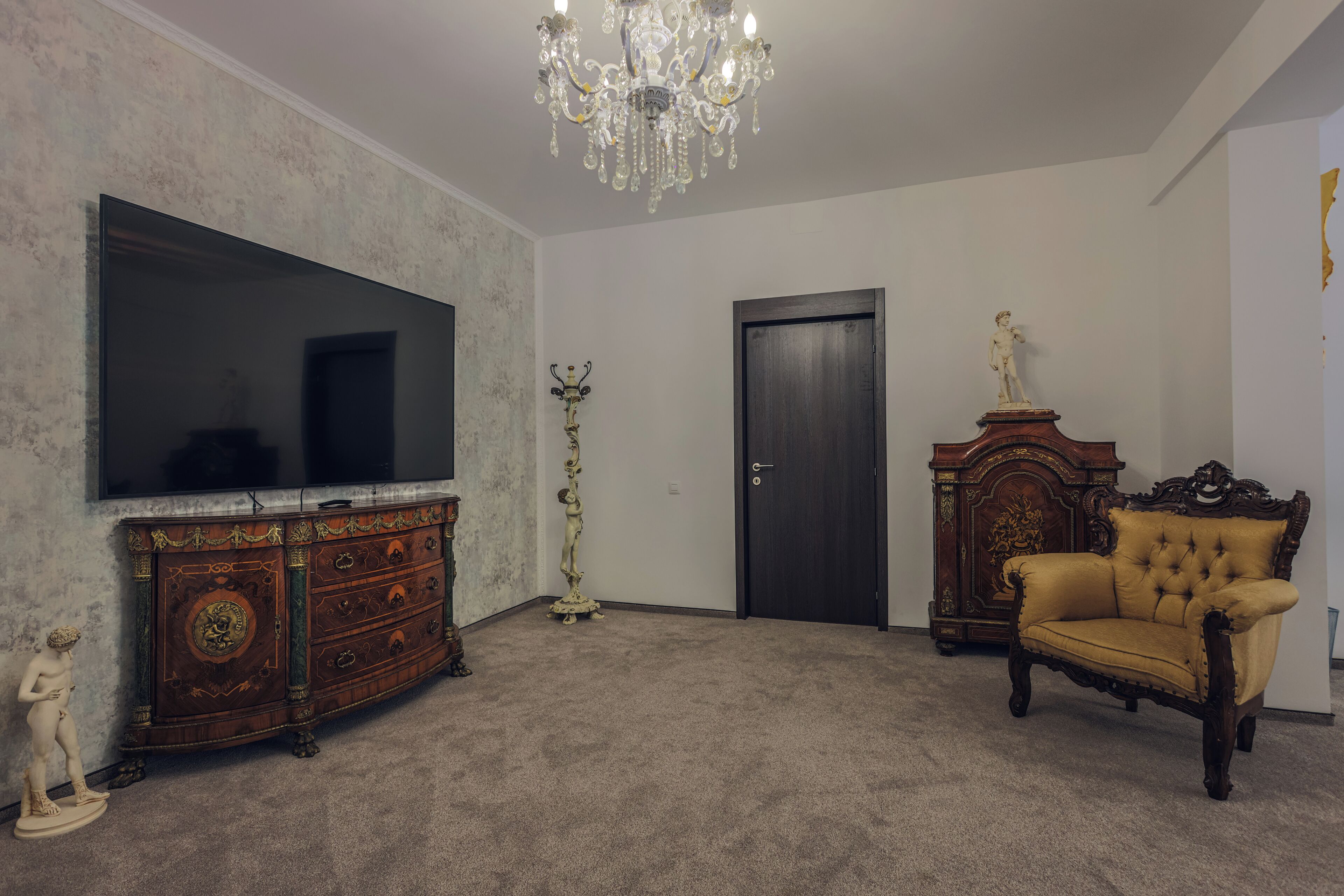 Een kamer met een grote flatscreen televisie, een houten kast, een hoge vloerlamp, een deur en een gestofte fauteuil.