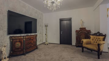 A room with a large flat-screen TV, a wooden cabinet, a tall floor lamp, a door, and a tufted armchair.