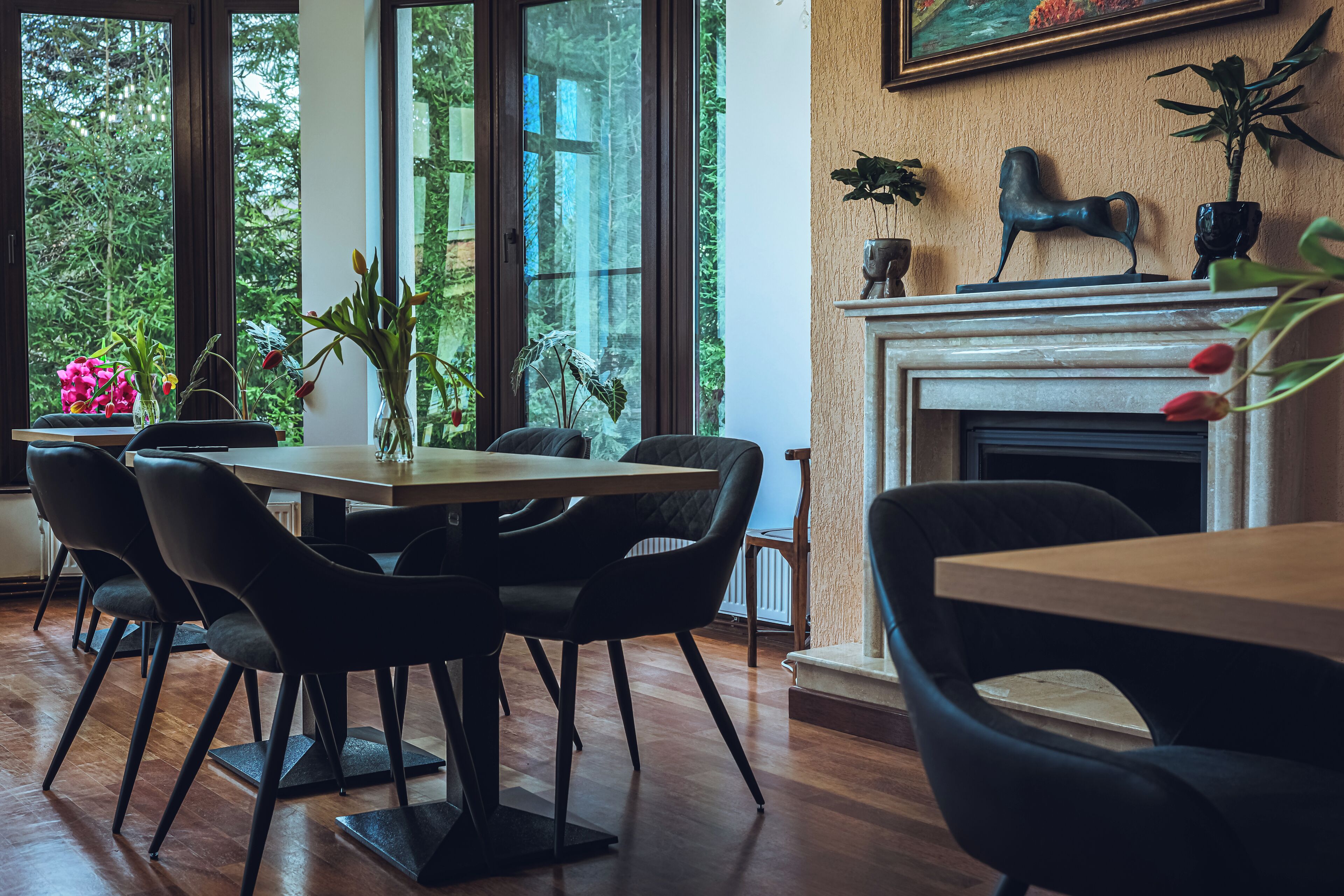 A dining area with a wooden table and chairs, a fireplace, and a view of greenery through large windows.