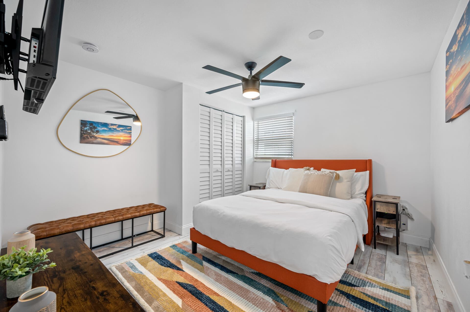 A bedroom with a bed, a ceiling fan, a bench, a bedside table, and a TV mounted on the wall.