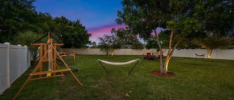 A backyard with a swing set, hammock, and trees.