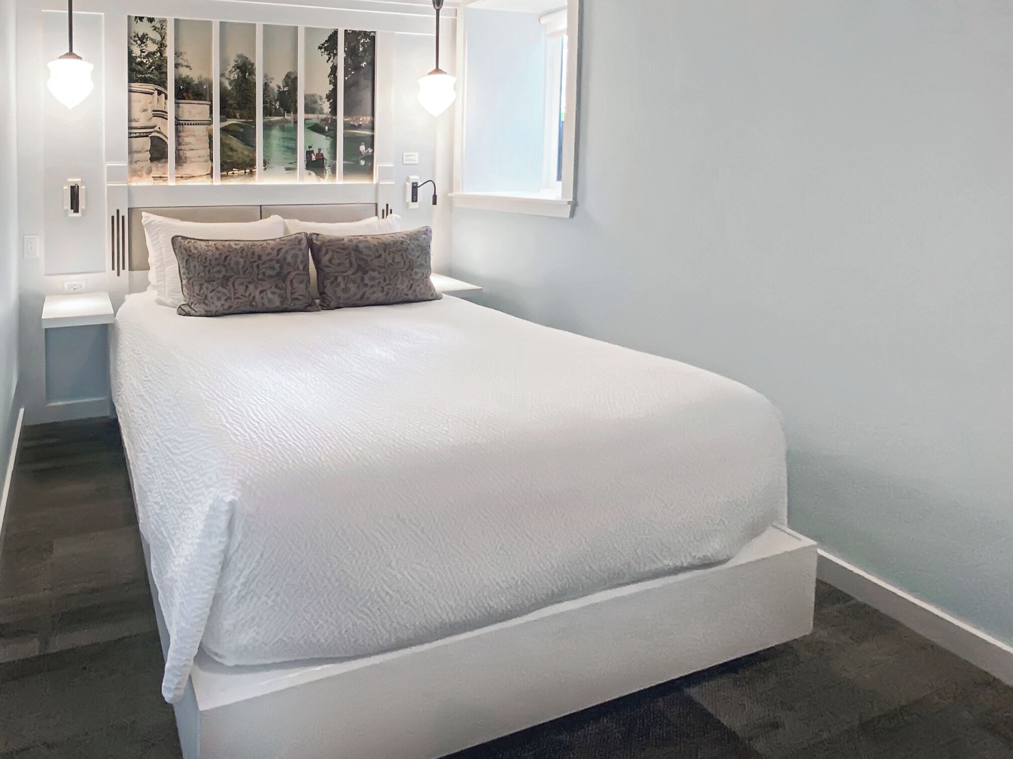 A bedroom with a large bed, a window with a view of a park, and two pendant lights.