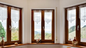 A dining room with a round table, wooden chairs, and a view of a mountainous landscape through large windows with decorative curtains. - Apartment in Kleines Wiesental (Kleines Wiesental - Raich)